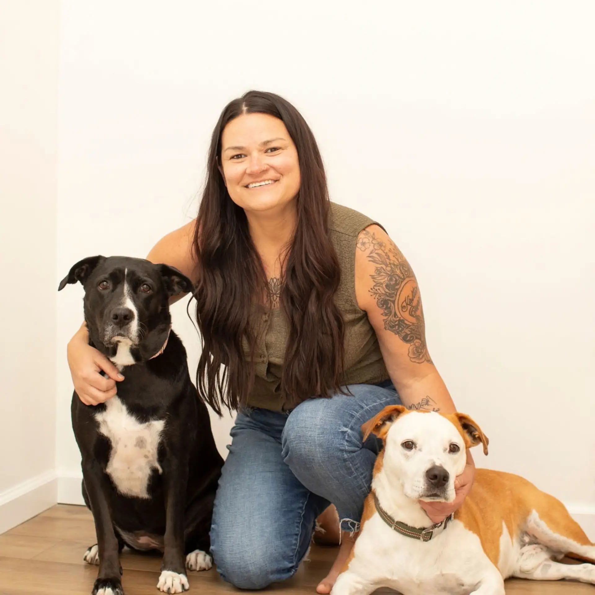 A woman with long dark hair sitting between two dogs; one white and brown on the right and a black dog on the left.